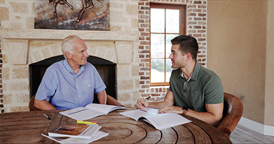 Dave Jewitt, sits at a wooden table next to a fireplace, and discusses Your One Degree with a partipant.