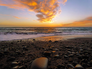 A view of the ocean from a rocky shore with the sun setting and reflecting on the water.
