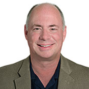 A headshot photo of Dr. Patrick Calhoun,&nbsp; wearing a olive green sportsjacket and a dark blue button up shirt.